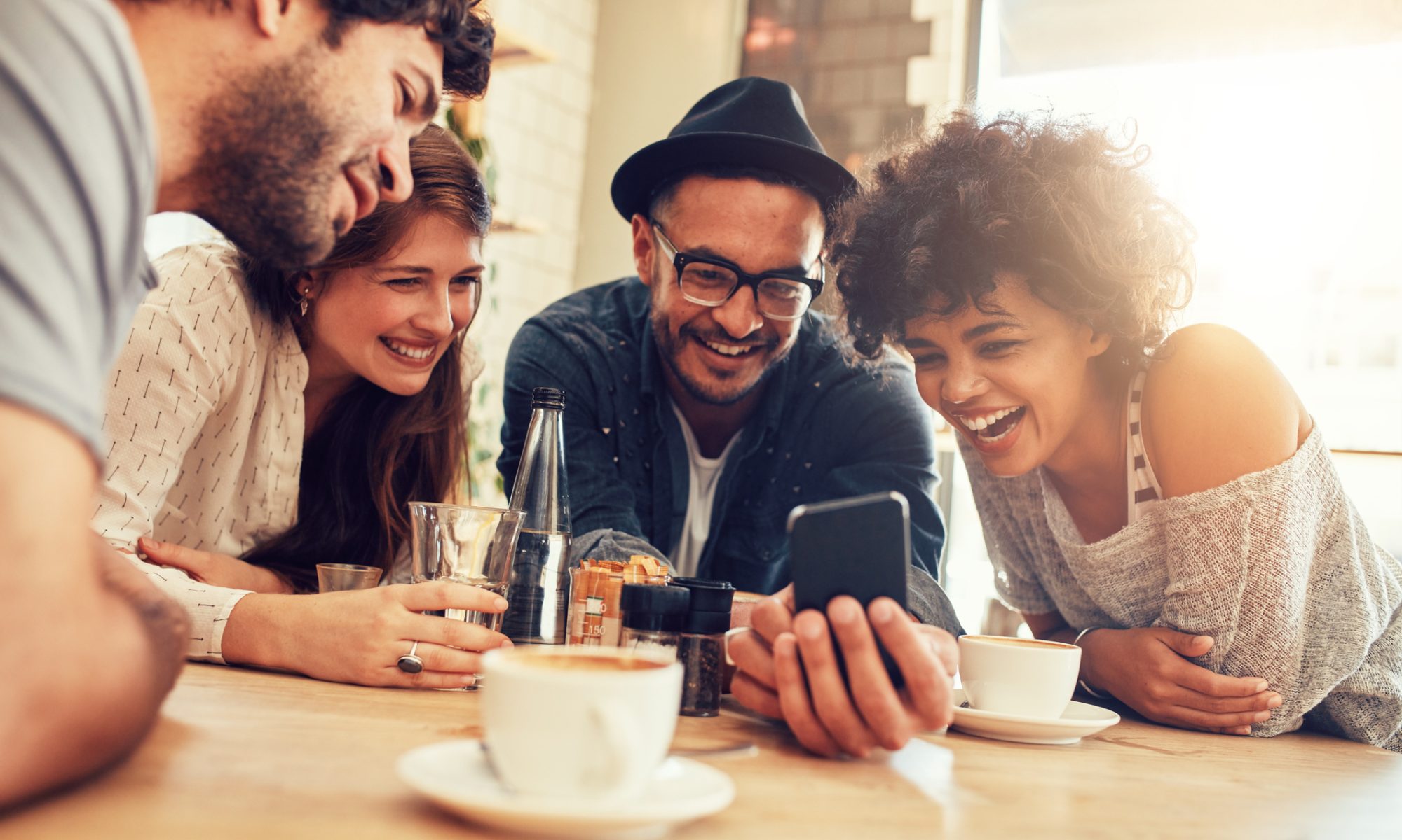 Portrait of cheerful young friends looking at smart phone while sitting in cafe. Mixed race people sitting at a table in restaurant using mobile phone.